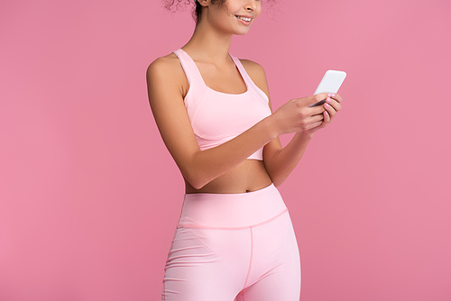 cropped view of happy young sportswoman using smartphone isolated on pink