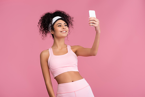 happy young sportswoman taking selfie on smartphone isolated on pink