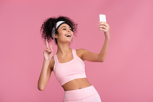 cheerful young sportswoman showing peace sign while taking selfie on smartphone isolated on pink