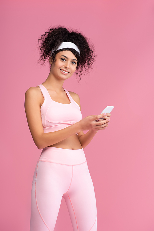 happy young sportswoman using smartphone isolated on pink