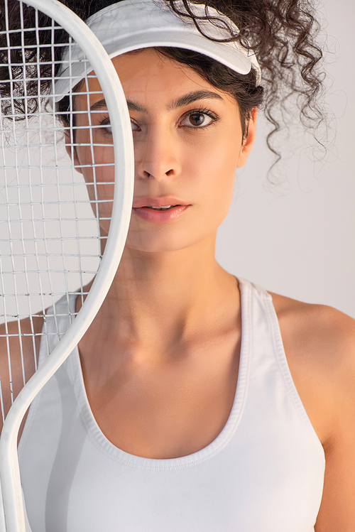 curly sportswoman in cap  through tennis racket isolated on white