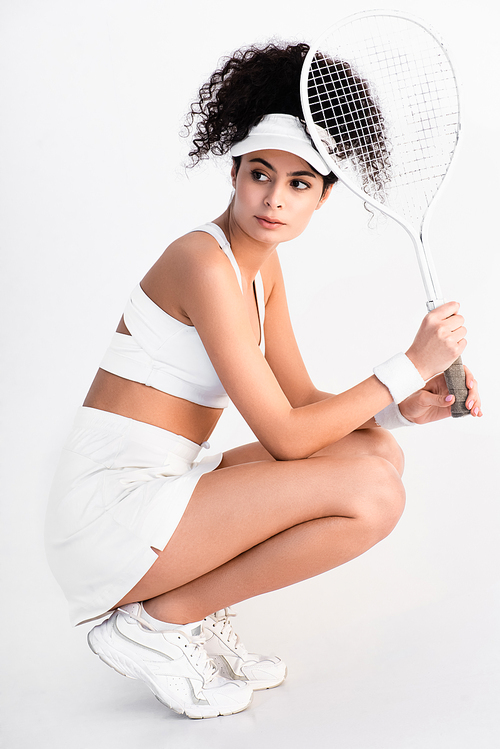 young sportswoman in cap sitting and holding tennis racket on white