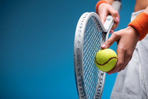 partial view of sportive young woman holding tennis racket and ball while playing on blue