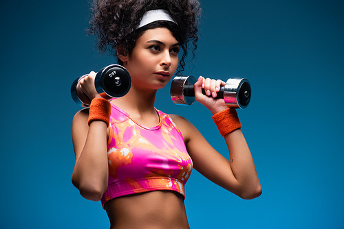 curly young sportswoman exercising with dumbbells on blue