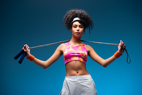 curly young sportswoman standing with skipping rope on blue