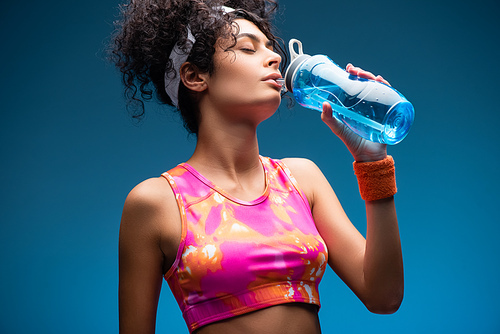 curly woman in sportswear drinking water while holding sports bottle on blue