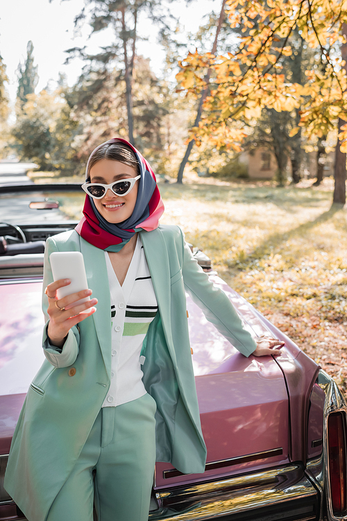 smiling, fashionable woman looking at smartphone while standing near vintage cabriolet