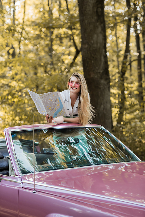 smiling woman in sunglasses looking at road atlas while sitting in cabriolet in forest