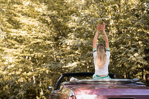 back view of young woman sitting in cabriolet with raised hands in forest