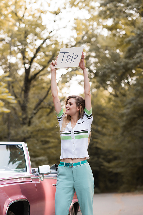 young woman holding placard with trip lettering in raised hands while standing near vintage convertible car