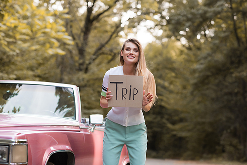 cheerful woman  while holding card with trip lettering near vintage convertible card