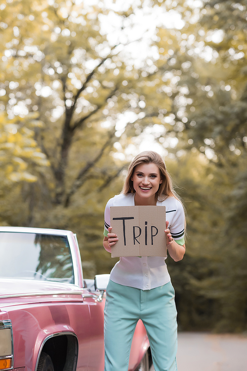 excited young woman holding placard with trip lettering near retro convertible car