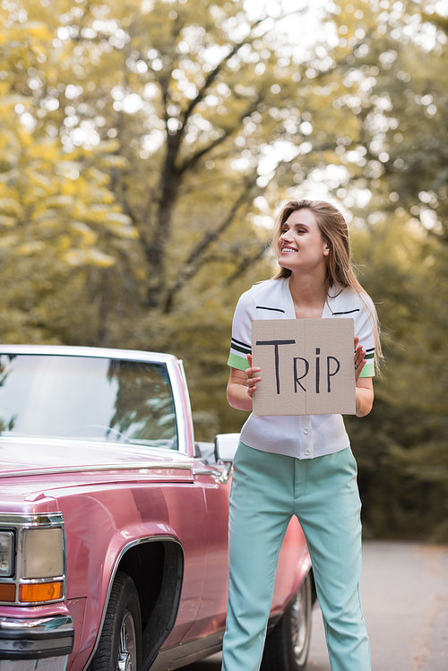 cheerful woman looking away while holding card with trip lettering near vintage convertible car