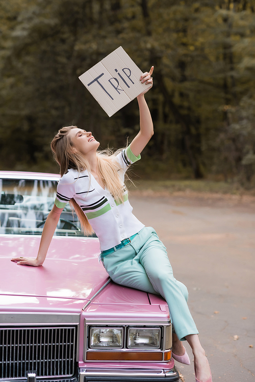 smiling woman holding placard with trip lettering while sitting on hood of cabriolet