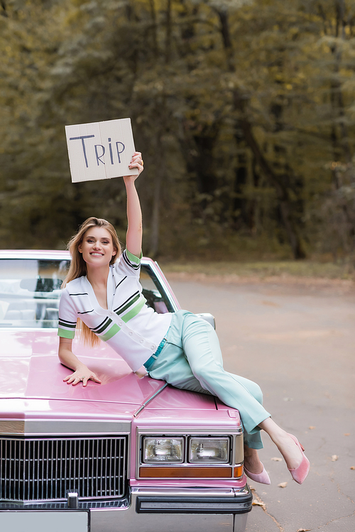 stylish young woman holding placard with trip lettering while lying on cabriolet hood