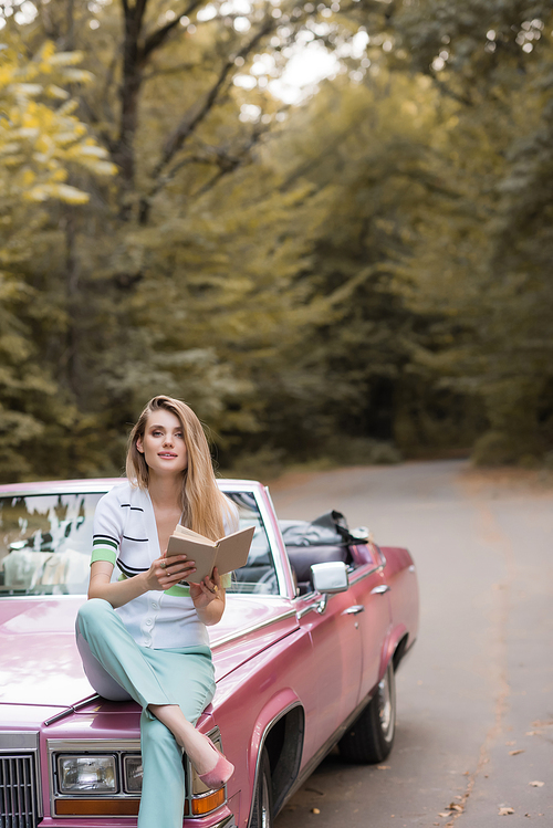 smiling woman  while sitting on hood of cabriolet and holding book