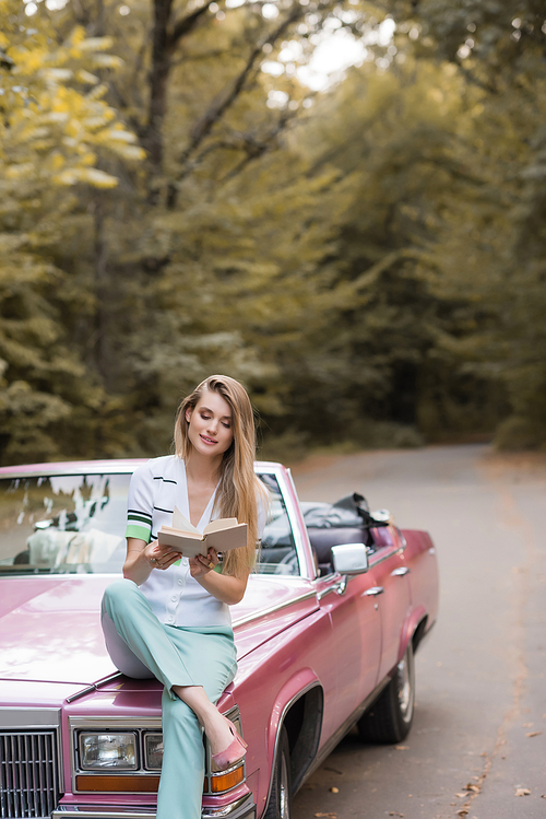 young smiling woman sitting on hood of vintage cabriolet and reading book
