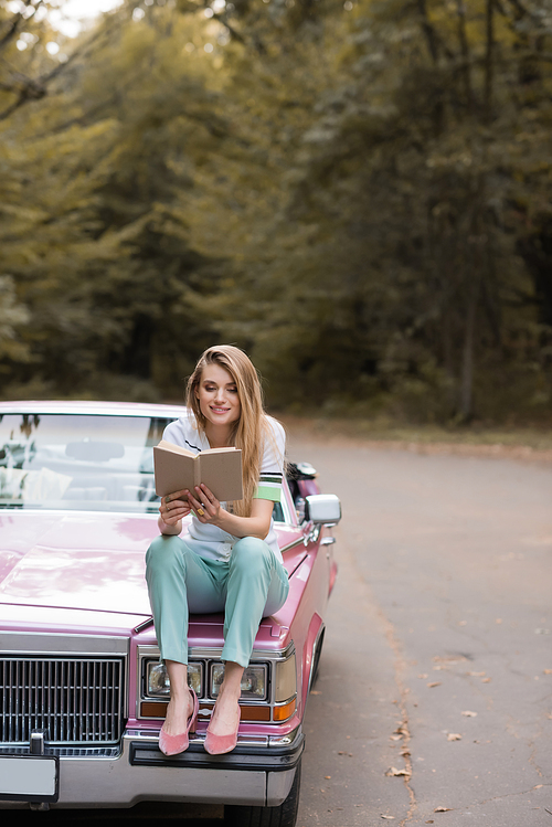 smiling woman sitting on hood of cabriolet and reading book