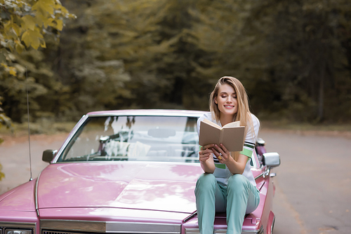 happy young woman reading book while sitting on hood of convertible car on blurred background