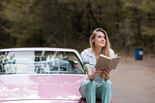 happy woman looking away while sitting on hood of cabriolet and looking away