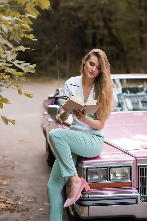 stylish woman reading book while sitting on hood of vintage cabriolet