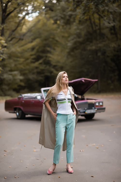 stylish woman with hands in pockets standing on road near broken cabriolet on blurred background