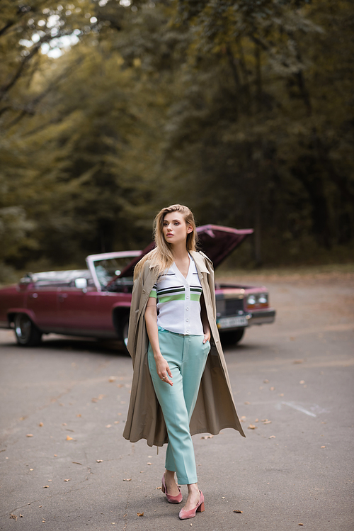 young stylish woman walking with hand in pocket near broken cabriolet with open hood on blurred background