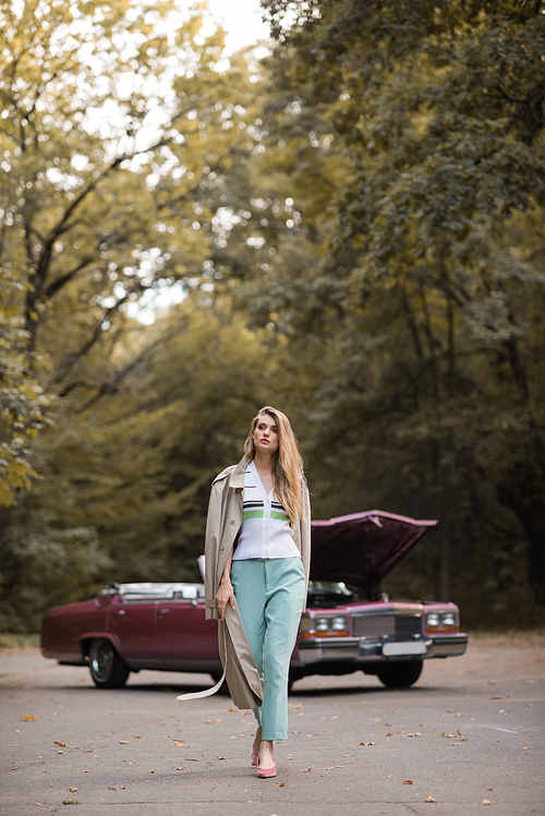 stylish woman in cape walking on road near broken vintage car on blurred background