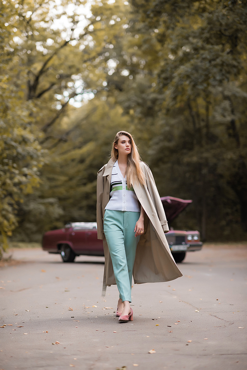 stylish woman in cape looking away while walking on road near broken retro car on blurred background