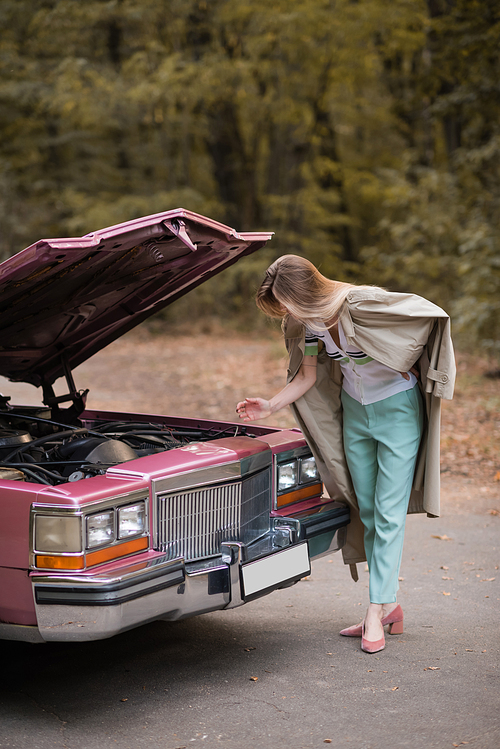 young woman looking under open hood of broken vintage car on road