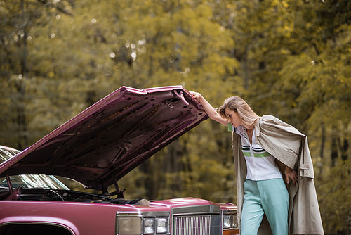 young woman in cape looking under open hood of broken vintage car