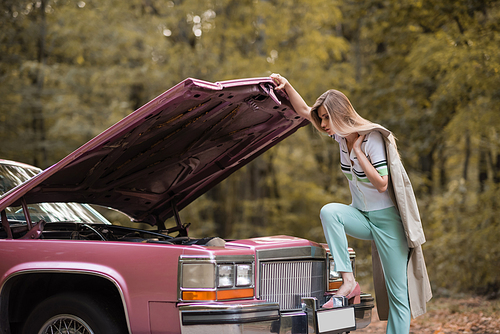 young stylish woman looking under open hood of broken retro car