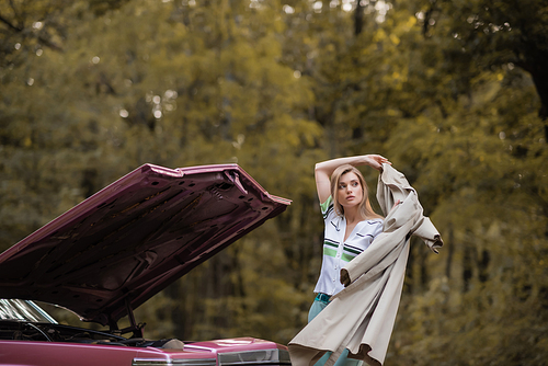 young woman putting cape on while standing near broken vintage car with open hood