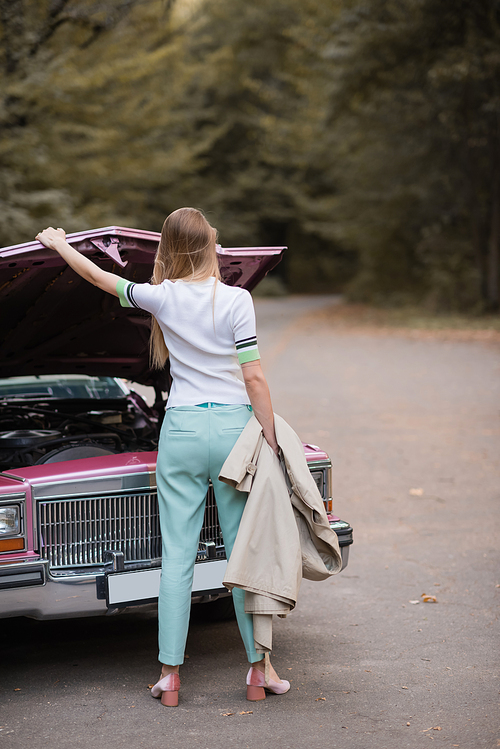 back view of young woman holding cape while opening hood of broken retro car