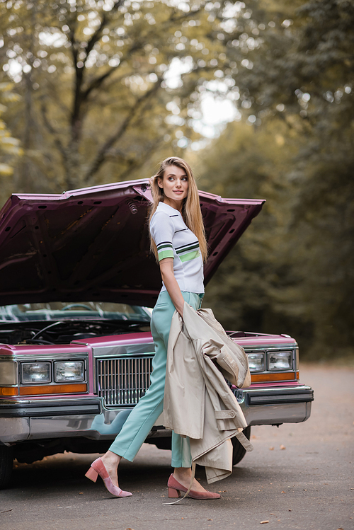 stylish woman holding cape and looking away while opening hood of broken retro car