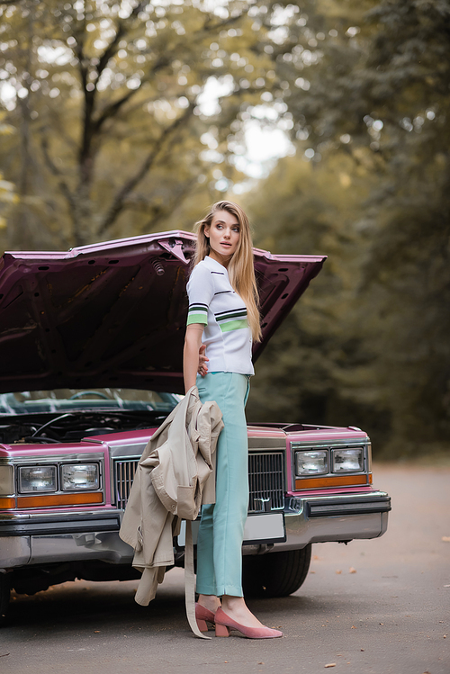 young woman holding cape and looking away while opening hood of broken vintage car