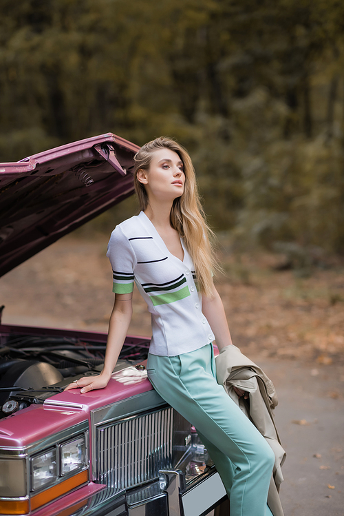young stylish woman looking away while standing near broken retro car with open hood