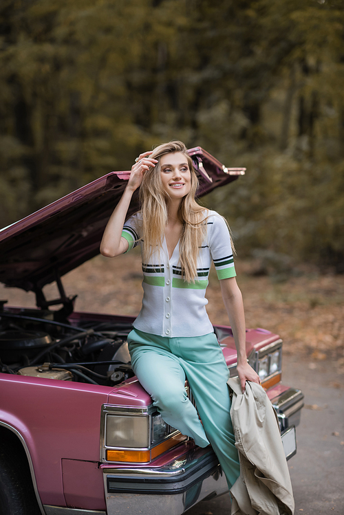 smiling woman touching hair and looking away while sitting on broken car with open hood