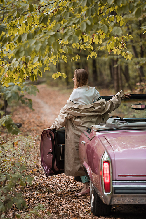 back view of woman in cape standing near open door of cabriolet in forest and looking away