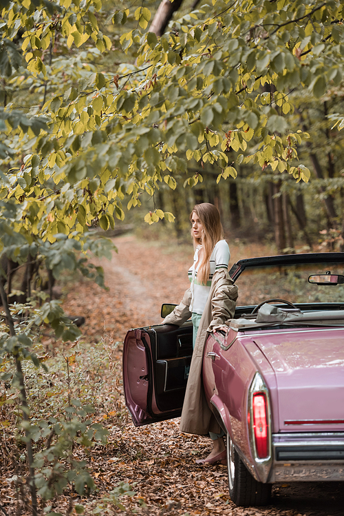 stylish woman looking away while standing near open door of cabriolet in forest