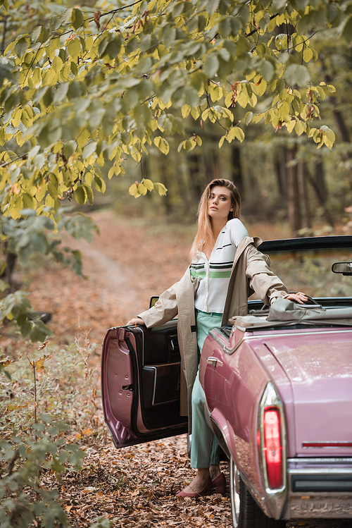 young stylish woman in cape  while standing near vintage cabriolet in forest