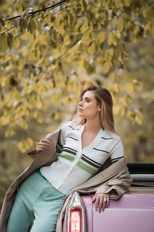 young woman in cape looking away while posing near cabriolet in forest