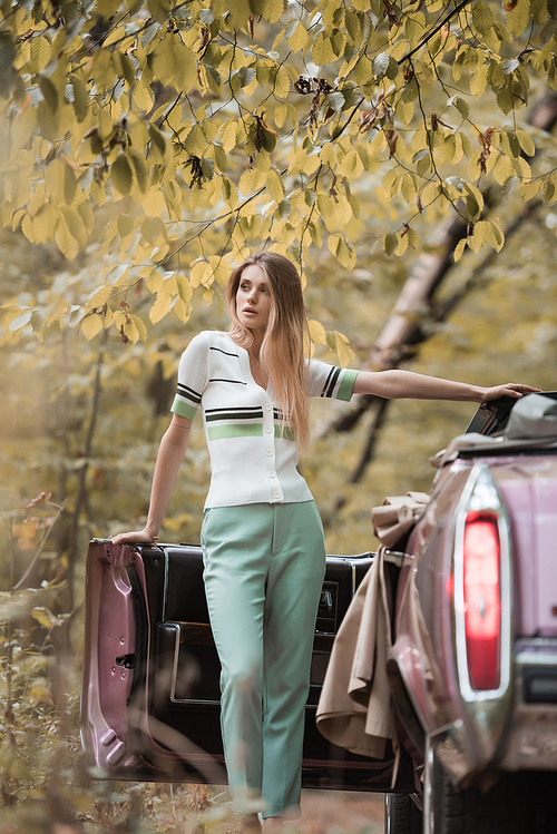 young woman looking away while standing near open door of cabriolet on blurred foreground