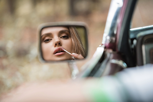 woman applying lip gloss while looking at side view mirror on blurred foreground