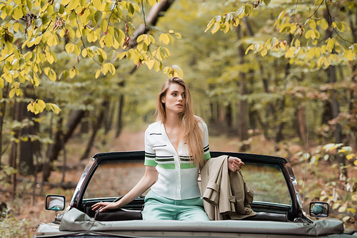 young woman holding cape while sitting in cabriolet and looking away in forest