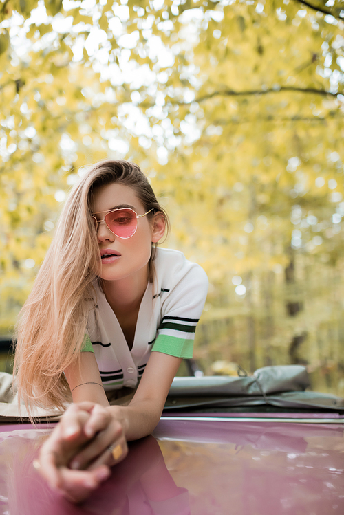 young stylish woman in sunglasses leaning on hood of cabriolet on blurred foreground