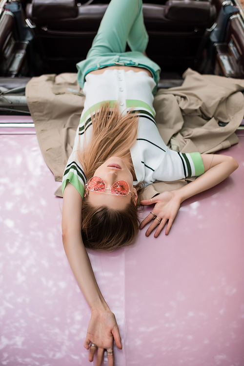 overhead view of stylish woman in sunglasses relaxing while lying on car on blurred background