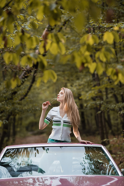 young woman holding sunglasses and looking away while standing in retro cabriolet