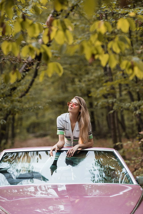 young woman in sunglasses looking away while leaning on windshield of convertible car