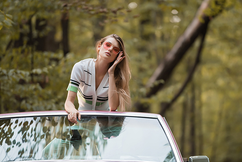 young woman touching sunglasses while leaning on windshield of convertible car